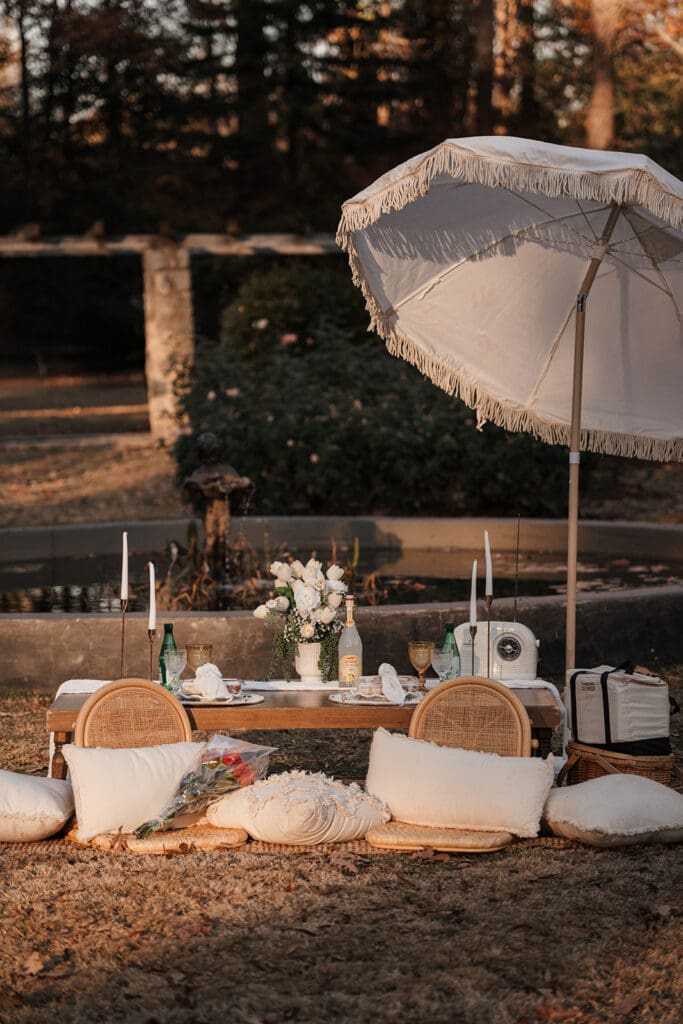 Elegant luxury picnic setup at Raleigh Rose Garden proposal with umbrella, candles, and florals arranged beside the fountain.
