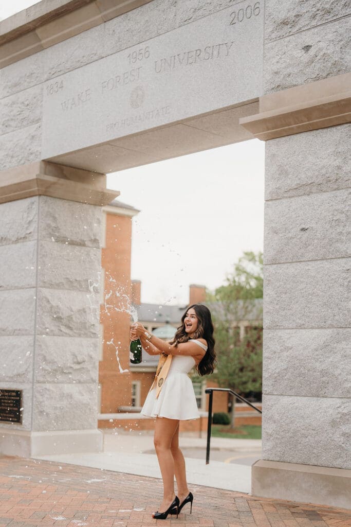 Graduate celebrating with champagne in front of stone archway on Wake Forest University campus